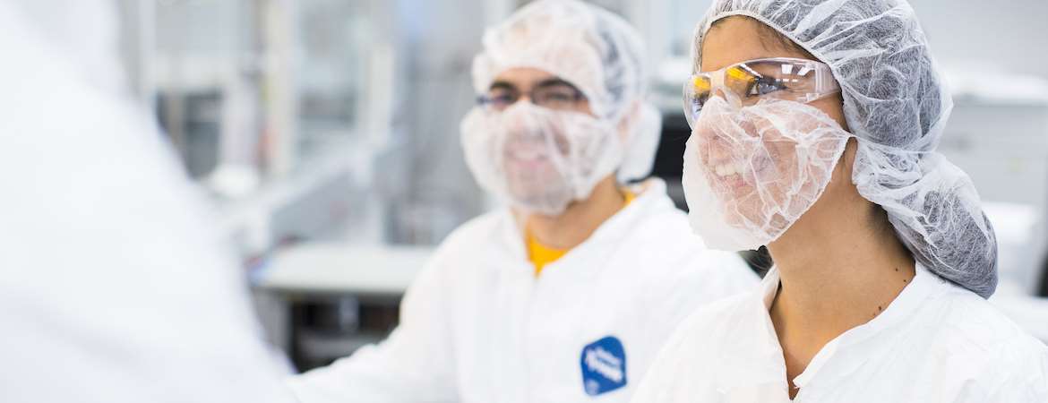 Two scientists wearing protective hair nets, face masks, goggles, and lab coats converse with another scientist who is only partially in-frame.